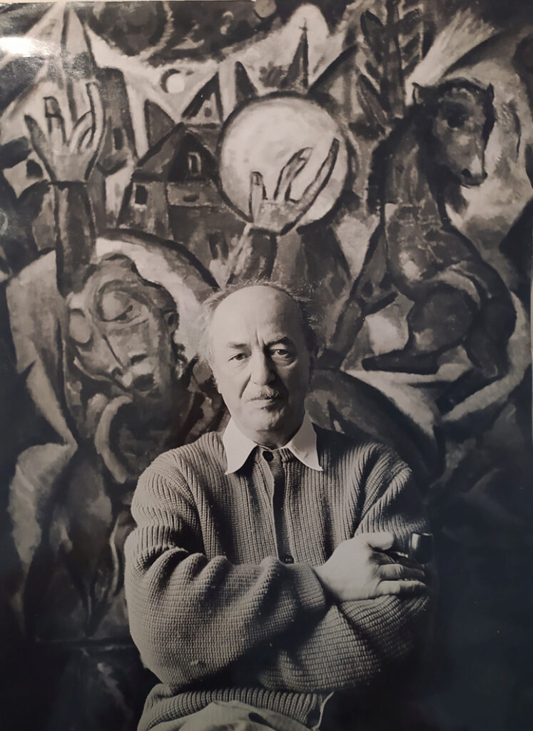 Schwarzweiss-Foto. Otto Gleichmann steht vor seinem Bild mit verschränkten Armen und Blickt in die Kamera. In der rechten Hand hält er eine Tabakspfeife.