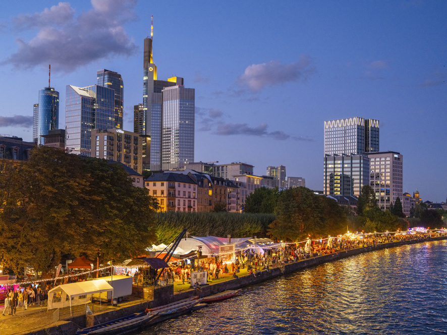 Frankfurt_Museumsuferfest 25_2099_©#visitfrankfurt_Holger_Ullma Aufnahme der Beleuchteten Buden am Frankfurter Mainufer in der Dämmerung. Im Hintergrund die Hochhäuser der Frankfurter Skyline.