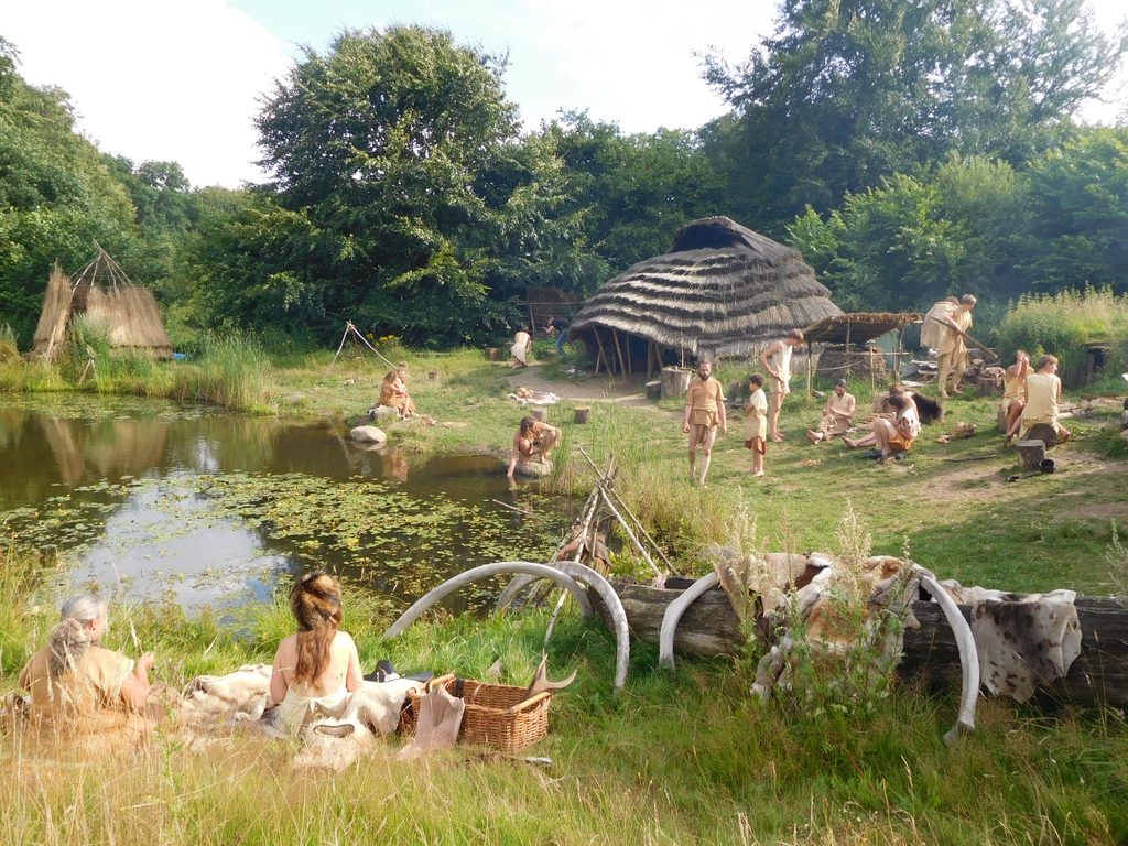An einem See lagern steinzeitlich gekleidete Menschen. Sie Bereiten Essen vor, arbeiten handwerklich und unterhalten sich. Im Hintergrund eine Schilf-Gedeckte offene Hütte