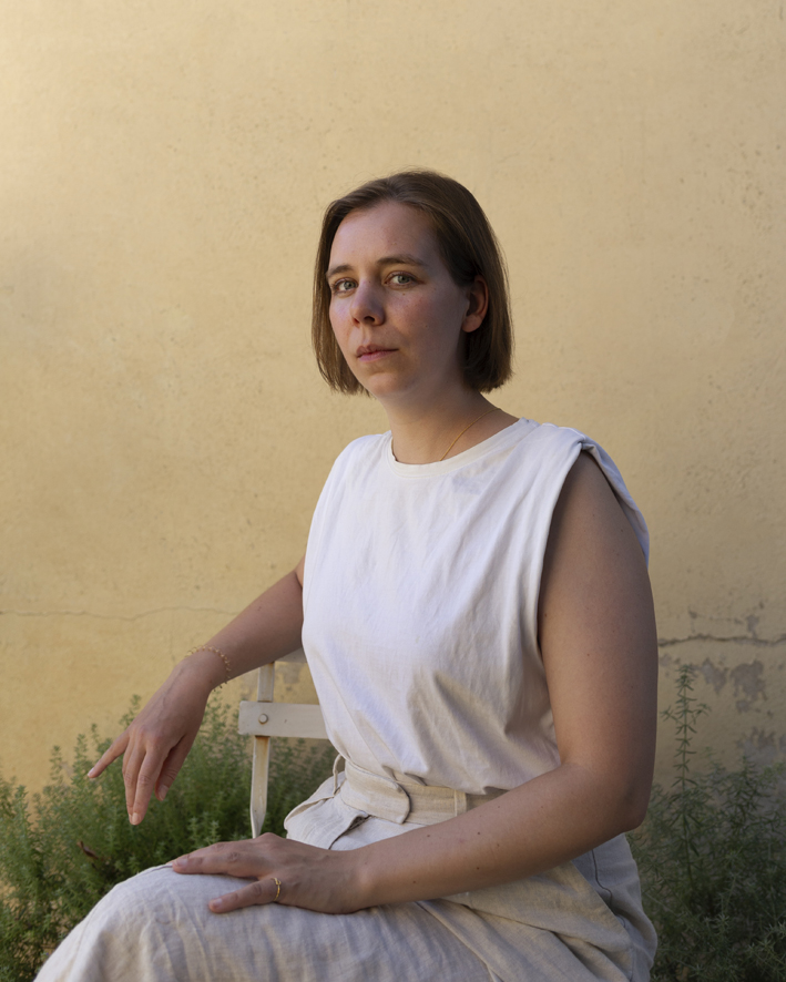 Clara Bolin sitzend auf einem Gartenstuhl. Der rechte Arm auf der lehne. Blick in die Kamera.