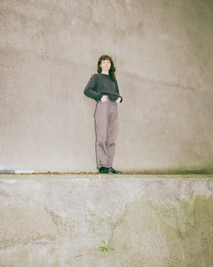 Frau steht vor einer grauen wand.Sie blickt in die Kamera, die Hände in den Taschen.